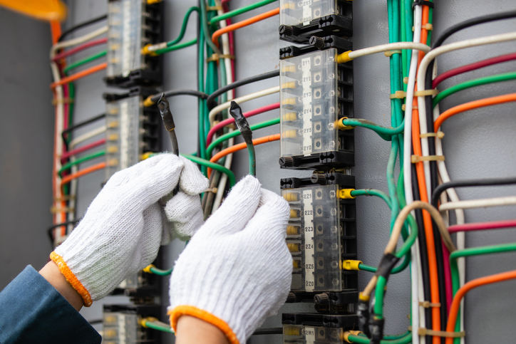 Electrical technician working on circuit board connections with colorful wires in control panel, wearing protective gloves and focusing on tasks